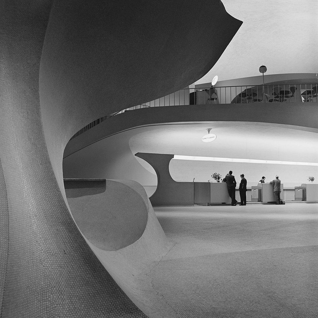 In the lobby of an airport building, there is a reception desk with two men standing next to it and several flight waters on display tables in front of them. The ceiling features curved curves and large windows, designed by [Eero Saarinen](https://goo.gl/search?artist%20Eero%20Saarinen). Blackandwhite photos, architectural photography, architectural design, curved shapes, circular space composition, and concrete walls. Aerial view perspective, high contrast, cool tones, and symmetrical layout.,,in