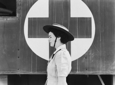 A black and white photograph of an Asian nurse standing in front of the side view profile of her medical train car, with "X" painted on it. She is wearing vintage fashion from around world war two era, including a hat. The background features details such as a cross symbol, solid color background, and a patterned border that adds depth to the scene. Shot in the style of [Dorothea Lange](https://goo.gl/search?artist%20Dorothea%20Lange).