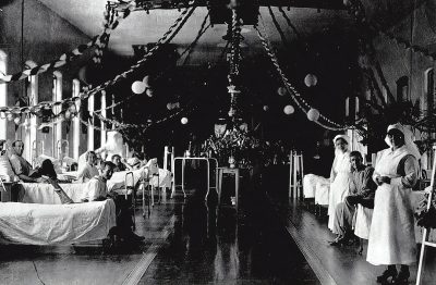 A black and white photograph of the interior of an old hospital room decorated for Christmas, with patients in beds and nurses wearing vintage attire. The scene captures festive decorations like garlands hanging from the ceiling and colorful paper balls hung on chair backs and table corners, all creating a warm atmosphere during the Christmas time. In one corner sits an elderly woman holding her hands together near her chest area. High resolution, in the style of an early 20th century photographer.