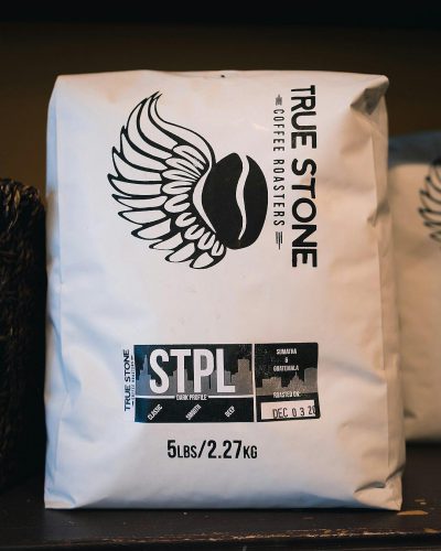 A large white coffee bag with the words "True Stone Coffee Roogoers" and logo of wings, black text that says "STpteL", on it is also written in smaller font, ‘5pants’ under '2 Sulit Ch preparing weight from Hey Foreigner', add an icon for water, below should be text reading, ‘4 LINFboard'. The design has a sleek modern look with clean lines and bold typography, conveying elegance through its simplicity and sophistication.