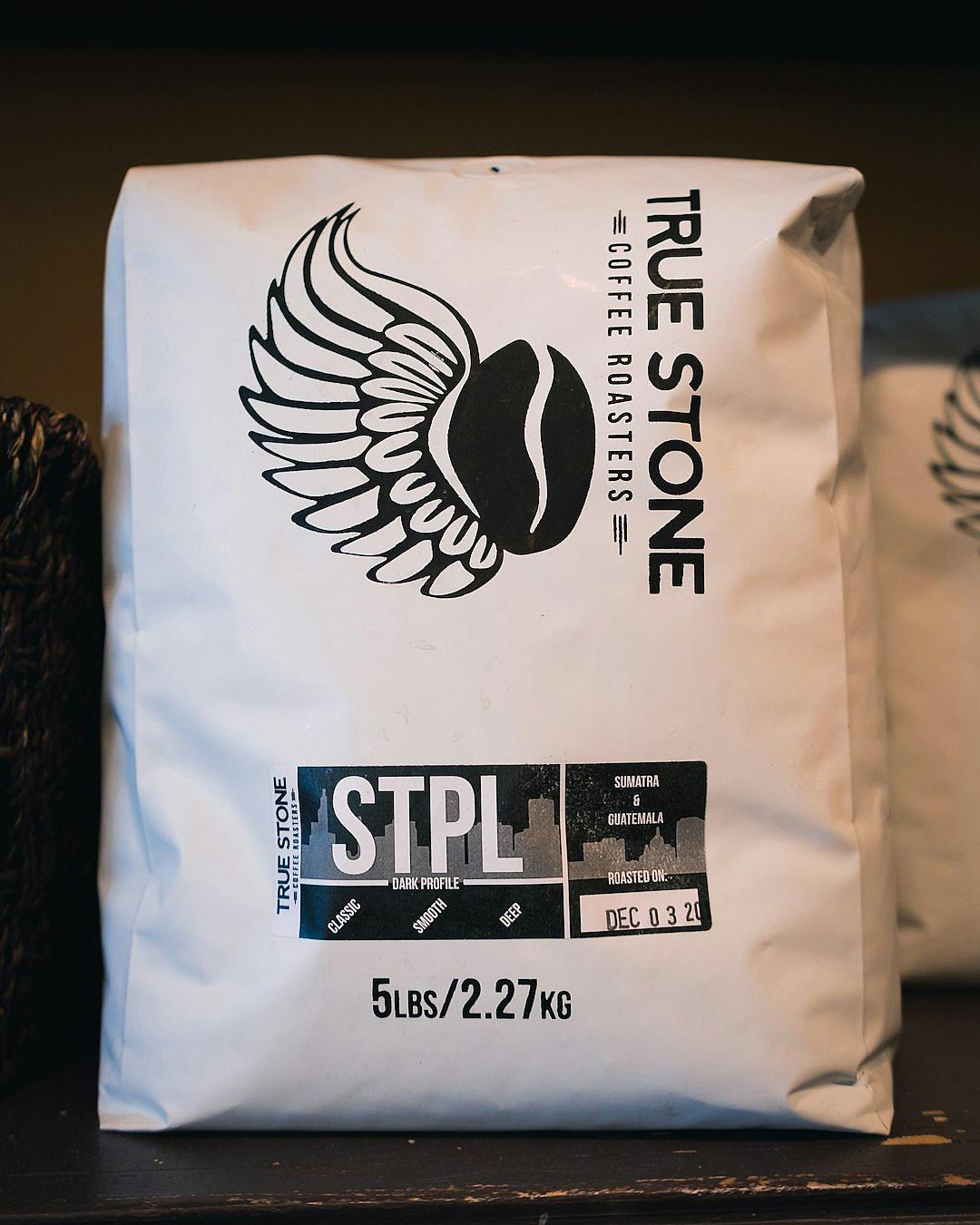 A large white coffee bag with the words “True Stone Coffee Roogoers” and logo of wings, black text that says “STpteL”, on it is also written in smaller font, ‘5pants’ under ‘2 Sulit Ch preparing weight from Hey Foreigner’, add an icon for water, below should be text reading, ‘4 LINFboard’. The design has a sleek modern look with clean lines and bold typography, conveying elegance through its simplicity and sophistication.