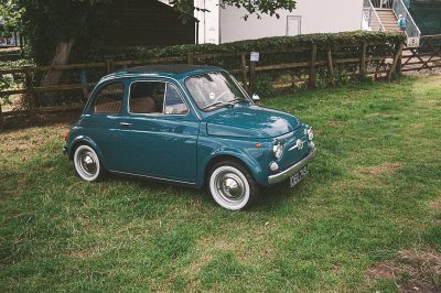 A blue Fiat 500 in the style of Leica film, with the look of Fujifilm Provia400F, in an outdoor grassy area resembling an old English village.