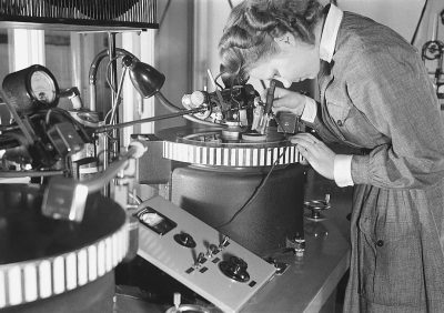 A woman is working on an audio tape recorder in the laboratory of electronic equipment, which was built by Jeltens at T Penrose in London in early monochrome photography. The main object she works with it is inside its wooden case and has four small rings around it that could be used to hold it down. It's set up on top of her desk where there’s also another two large tape recorders sitting next to each other. One of them has three buttons attached on one side.