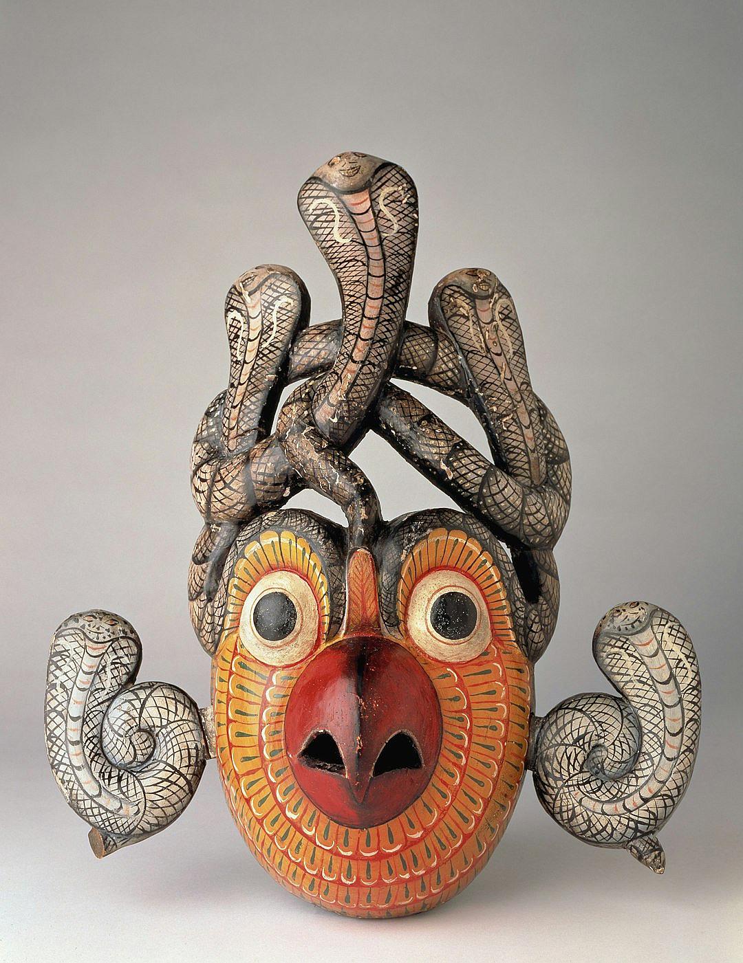 A wooden mask with snakes around the neck, inspired in the style of M.UPs and designed in folk art styles. It features an orange face, two eyes on each side of its head, dark red lips, four long tentacles wrapped around its mouth area, a small black nose at the center, multiple white scales covering the body, and six large gray arms hanging down from the shoulders. The background is a plain grey, creating contrast between lightness and darkness.