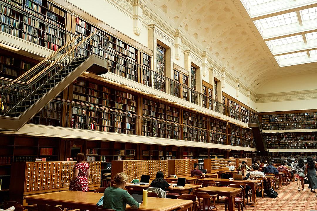 The State Library of Victoria in baby pink and dark brown, the grand reading room is filled with people studying books at tables. The walls have bookshelves stacked full of leather bound tomes from all eras, large skylights allowing natural light into the space.