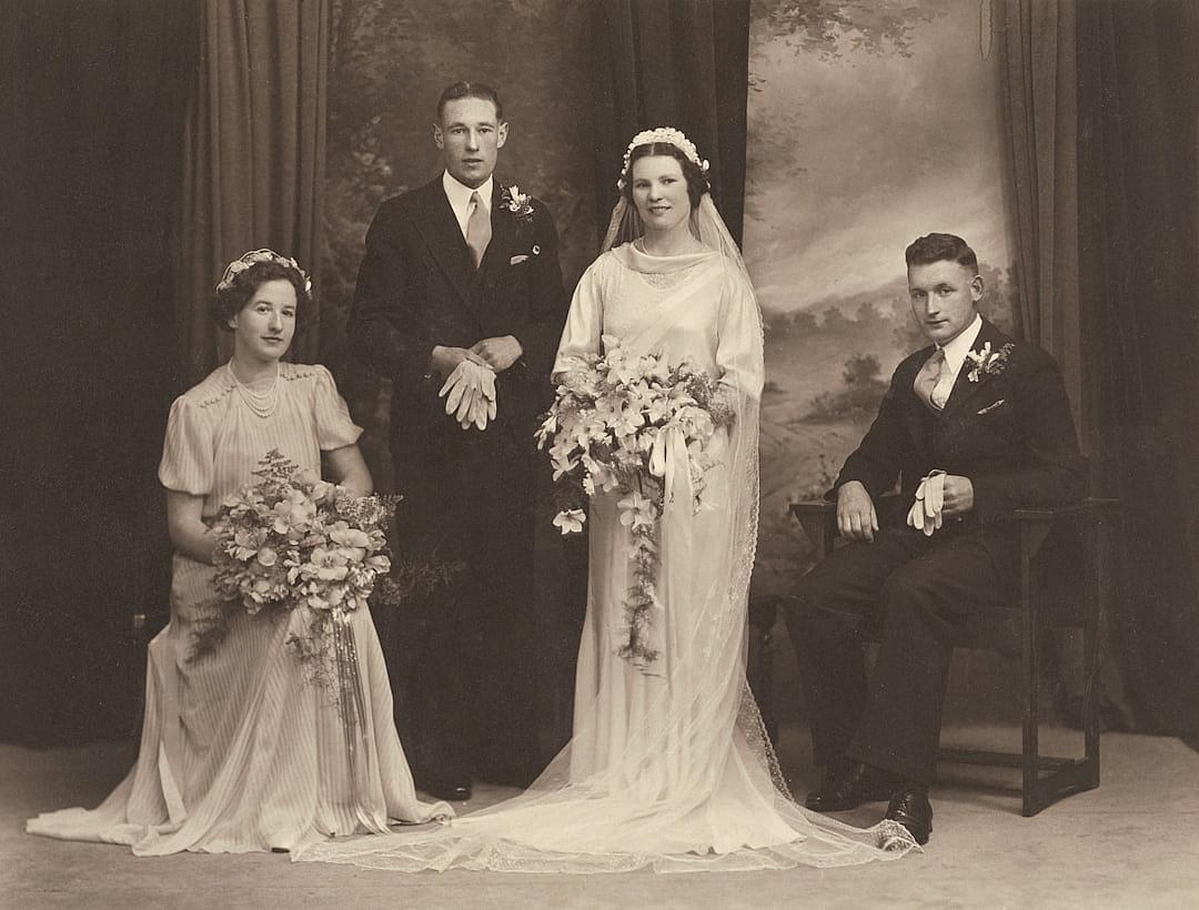 A vintage wedding photo shows the bride and groom with their two friends, all dressed in period attire from the 1930s to early 1940s. The bride wears an elegant white dress with lace detailing on her sleeves and neckline. She has flowers in her hair and holds a bouquet of roses. Her father stands beside them as he poses for photos, while his wife sits nearby holding onto one corner of his suit jacket. They stand against a backdrop that features a painting or drawing of nature, such as trees or mountains, in the style of that era.