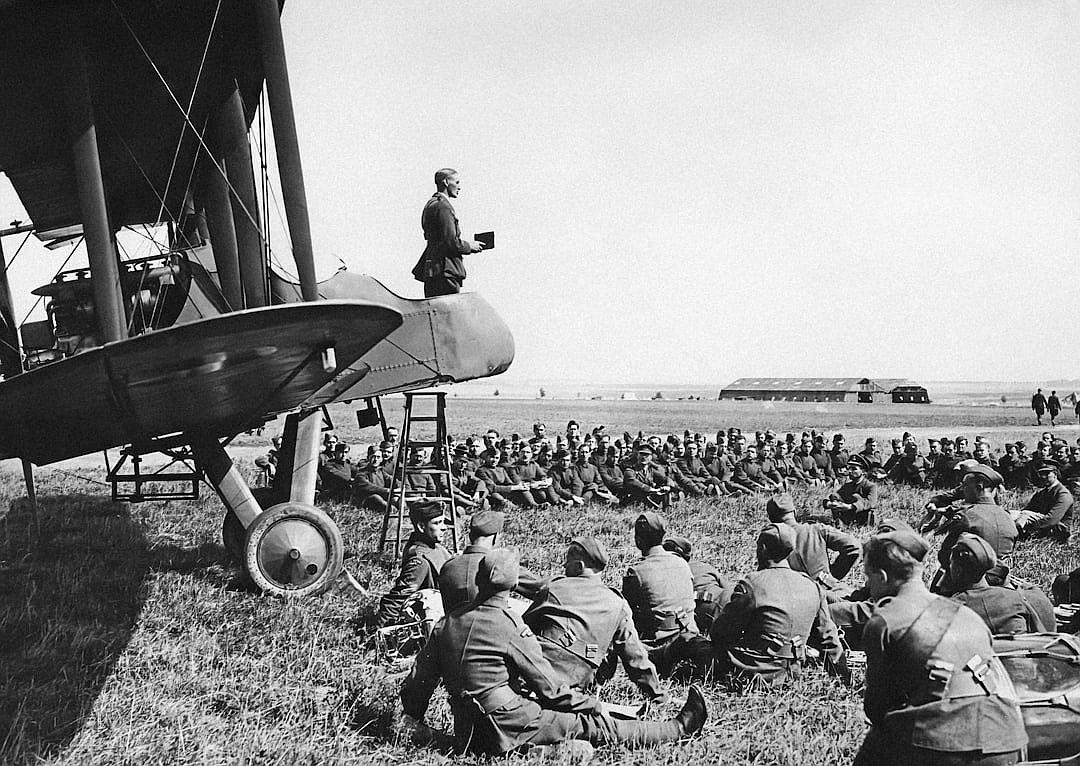 photo of German pilot standing on top of the biplane and giving a speech to soldiers sitting in a field, in the style of world war one era