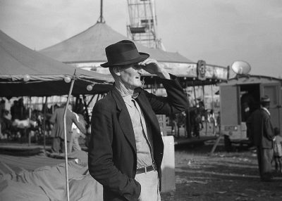 A black and white photograph of James Fordo, wearing his iconic hat and dark suit jacket with a light shirt underneath, standing in front of the circus tent on an empty fairground. He is looking off to one side while touching the brim of his fedora with his hand over his eye. In the background there are people working at market stalls. The photo was taken in the style of [Robert Capa](https://goo.gl/search?artist%20Robert%20Capa) using Kodak TriX film. It has a raw feel and features a shallow depth of field.