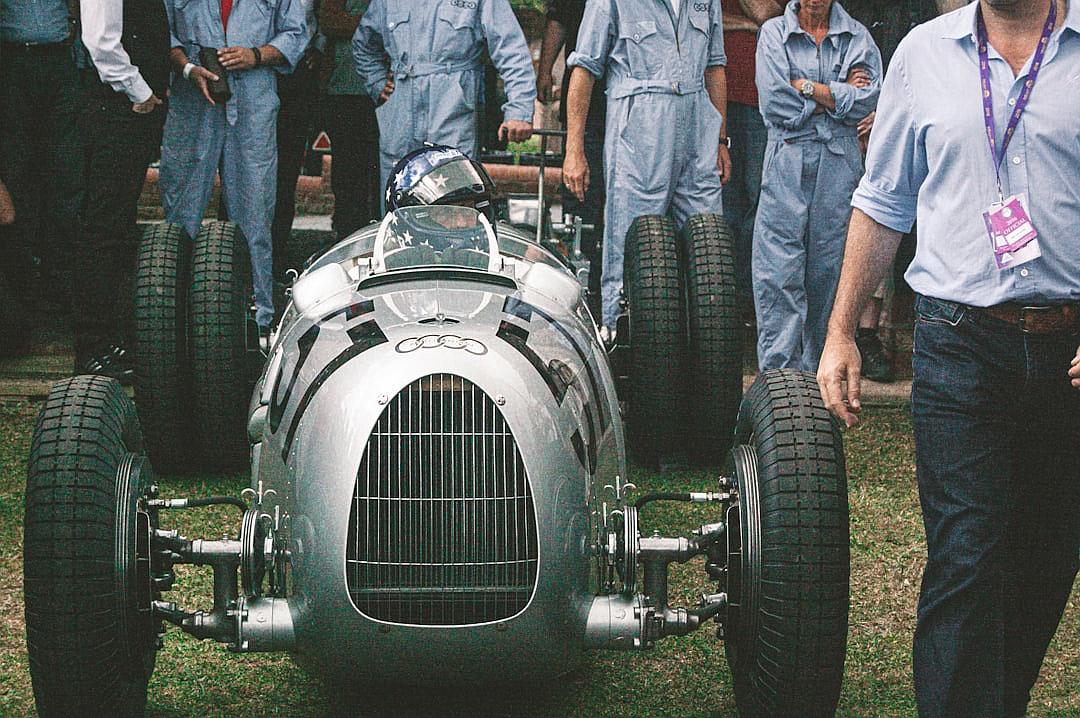 Close up of a vintage silver and grey art deco streamlined race car with wide tires. A front view shows the driver in a helmet standing next to the open hood on grass, surrounded by crew members wearing blue jumpsuits. Some are holding tools in bright daylight. Photographed in the style of a Leica M6 with a 50mm f/2 lens.