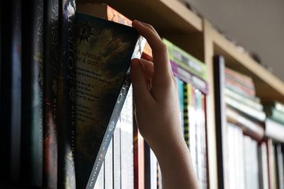 A hand reaches for the book "Akraine" on top of another shelf in between other books, with an illustration from one of its pages visible that shows an apocalyptic scene. The background is blurred and out of focus showing shelves filled with various fantasy-themed hardcover comics in the style of J. Rico or [Magali Villeneuve](https://goo.gl/search?artist%20Magali%20Villeneuve).