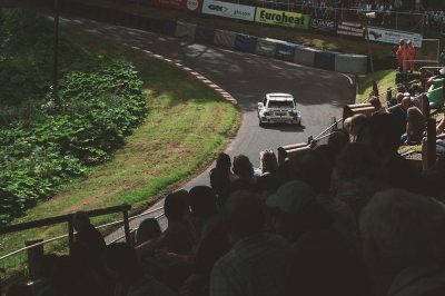 photo of an old rally car racing down the hill at high speed, there is some grass and trees on both sides of the track with people sitting in bleachers watching, the car has "esurveyorshopping.com" written on it's side. The crowd is cheering as they watch from their seats, shot by Hasselblad X2D camera