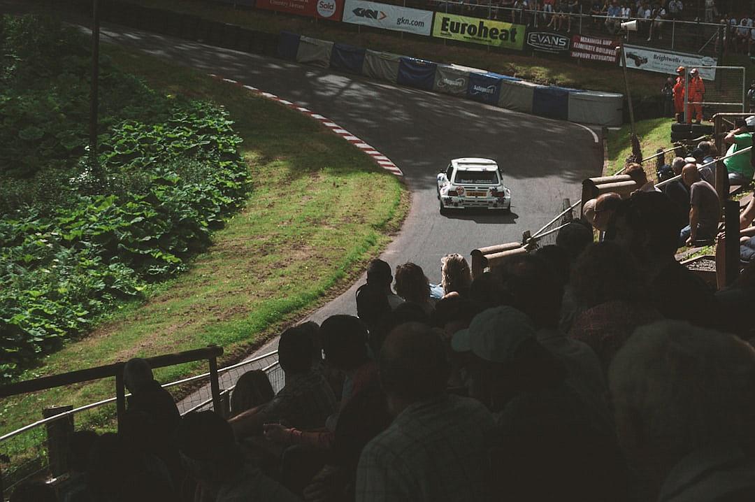 photo of an old rally car racing down the hill at high speed, there is some grass and trees on both sides of the track with people sitting in bleachers watching, the car has “esurveyorshopping.com” written on it’s side. The crowd is cheering as they watch from their seats, shot by Hasselblad X2D camera