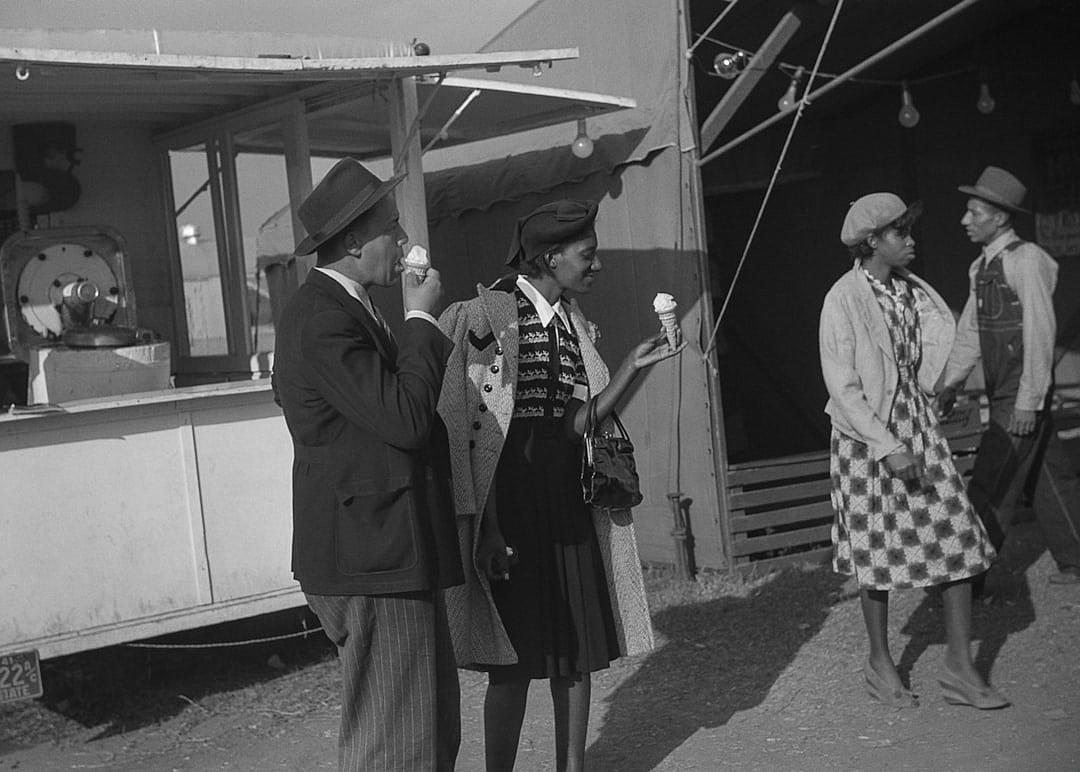 Black and white photo of African American people eating ice cream in front of an oldfashioned carnival food truck, mid20th century attire, retro style, lively atmosphere with a vintage feel, side view capturing the scene, Leica M3 camera with SummiluxM Griffiths lens, natural light