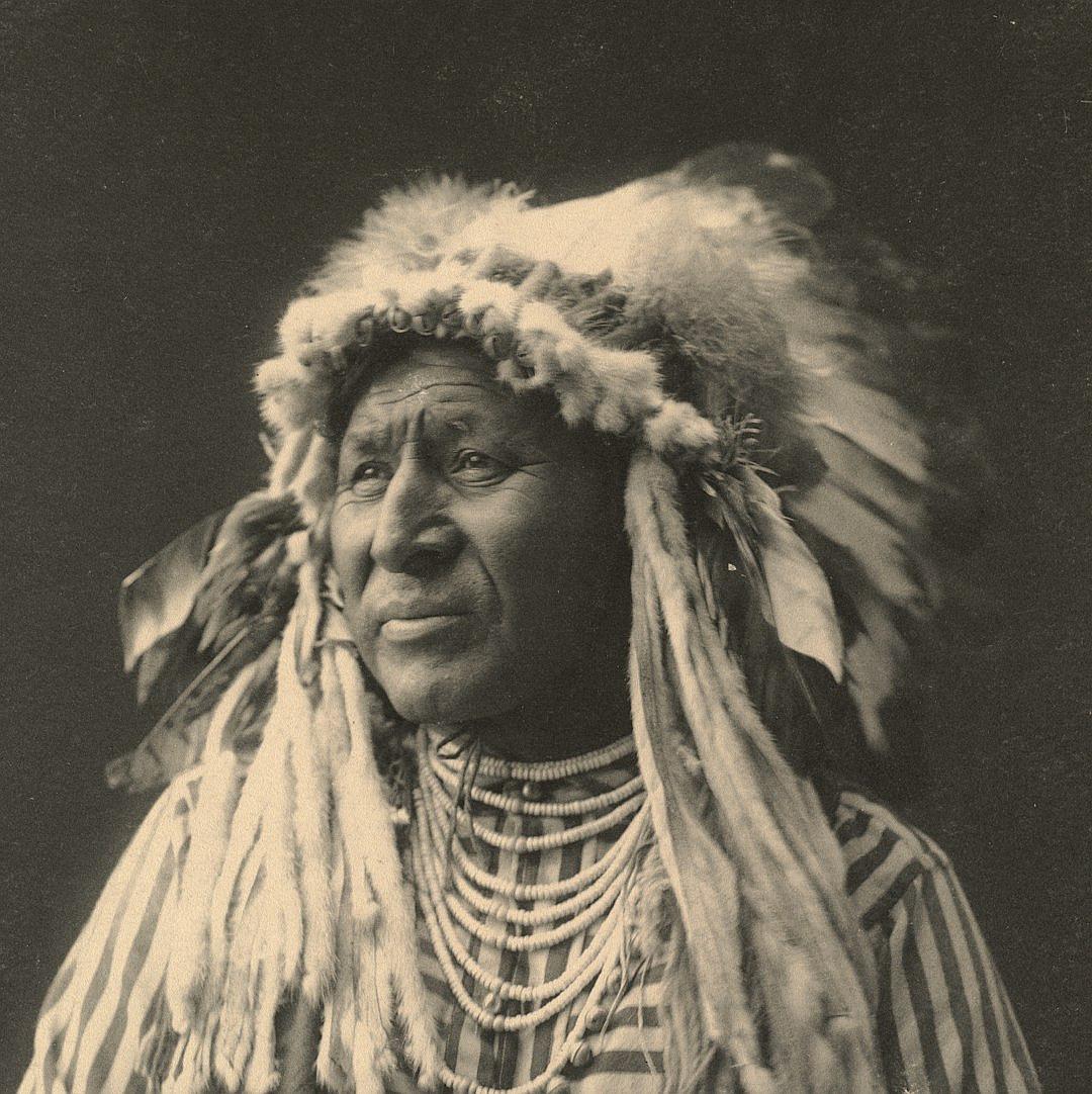 a vintage photograph of an Indian chief wearing his feathered headdress and striped robe, facing forward, 20th century photography