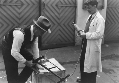 A black and white photo of two men in the street, one man is wearing an open vest with tie and hat holding a hair dryer, he stands next to another person who holds a metal device on which paper lies. They stand near each other, both looking at something. In front there's some kind of machine that has wires connected from it to a box lying nearby. The background features wooden doors. Shot in the style of Kary bridge.