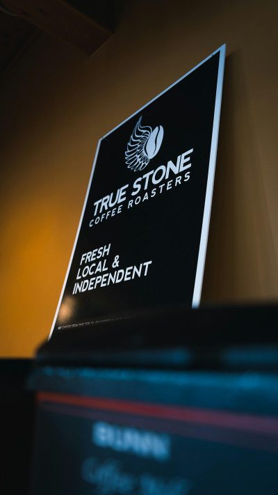 A closeup shot of an LED sign with the words "True Stone Coffee Roasters" on it, showcasing their logo and branding. The background is softly blurred to focus attention on the black coffee beans in front. A banner reads "Proudly serving local & independent". This photo captures True Stone's commitment to fresh roasting, urban style, and its position as one of the top independent coffee shops in the area. A shot of a private office at True Stone Coffee in Virginia, with warm lighting in the style of a minimalist design.