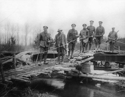 A black and white photo of British soldiers standing on a wooden bridge over a river, posing for a photograph with tools to build the bridge in hand, from the World War I era. The soldiers are posed in the style of early 20th century military photography.