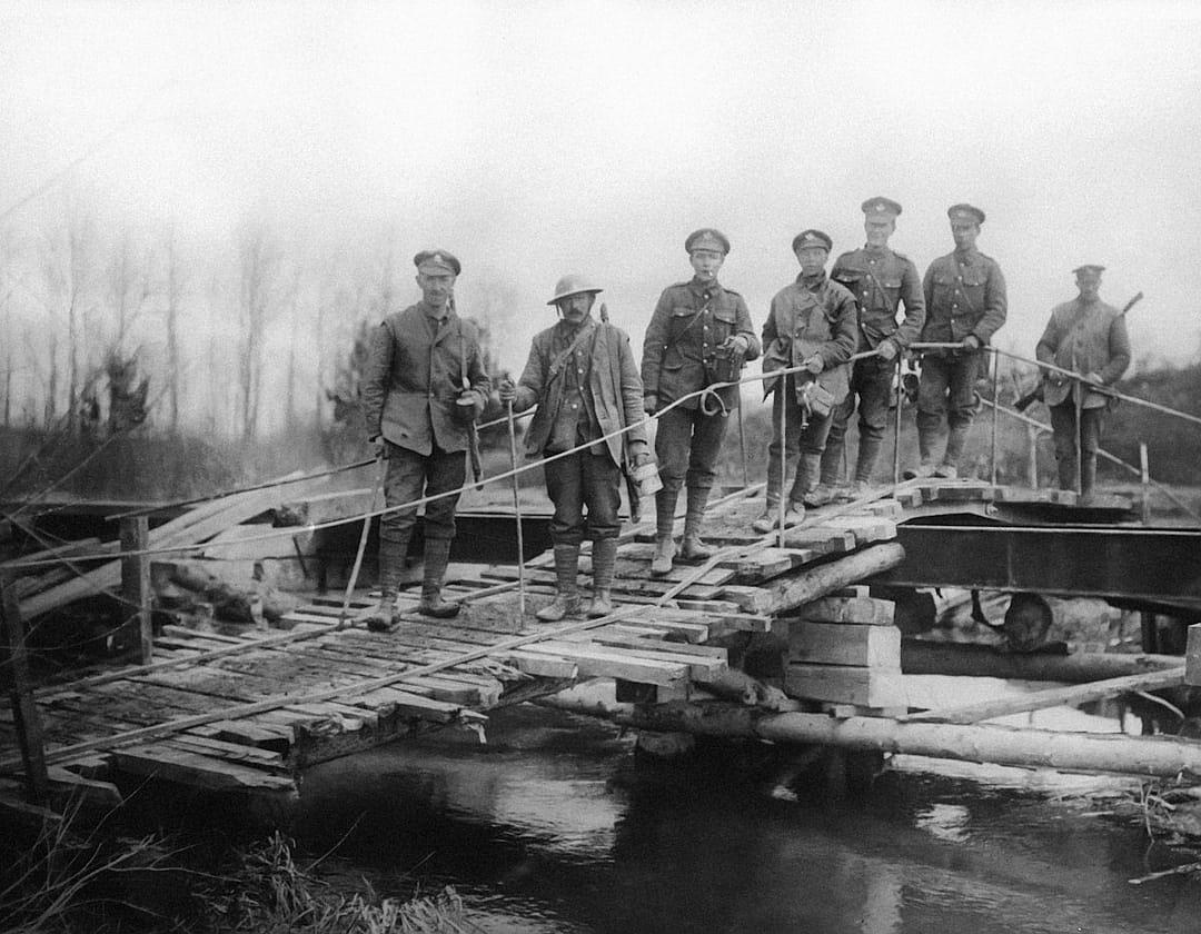 A black and white photo of British soldiers standing on a wooden bridge over a river, posing for a photograph with tools to build the bridge in hand, from the World War I era. The soldiers are posed in the style of early 20th century military photography.