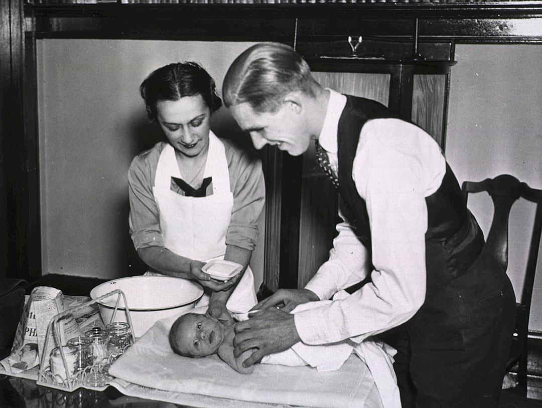 A black and white photo from the early 20th century shows a doctor performing the first paving. The scene includes two people, a man in his late thirties with short hair wearing formal attire including dark trousers and shoes, and a woman around twenty years old dressed as a nurse giving a baby monitor on a table covered with cloth. In front is a newborn lying down having its head patted with both hands, with medical equipment scattered across the surface of the tabletop. The background features wooden wall panels. The photography has a realistic style.