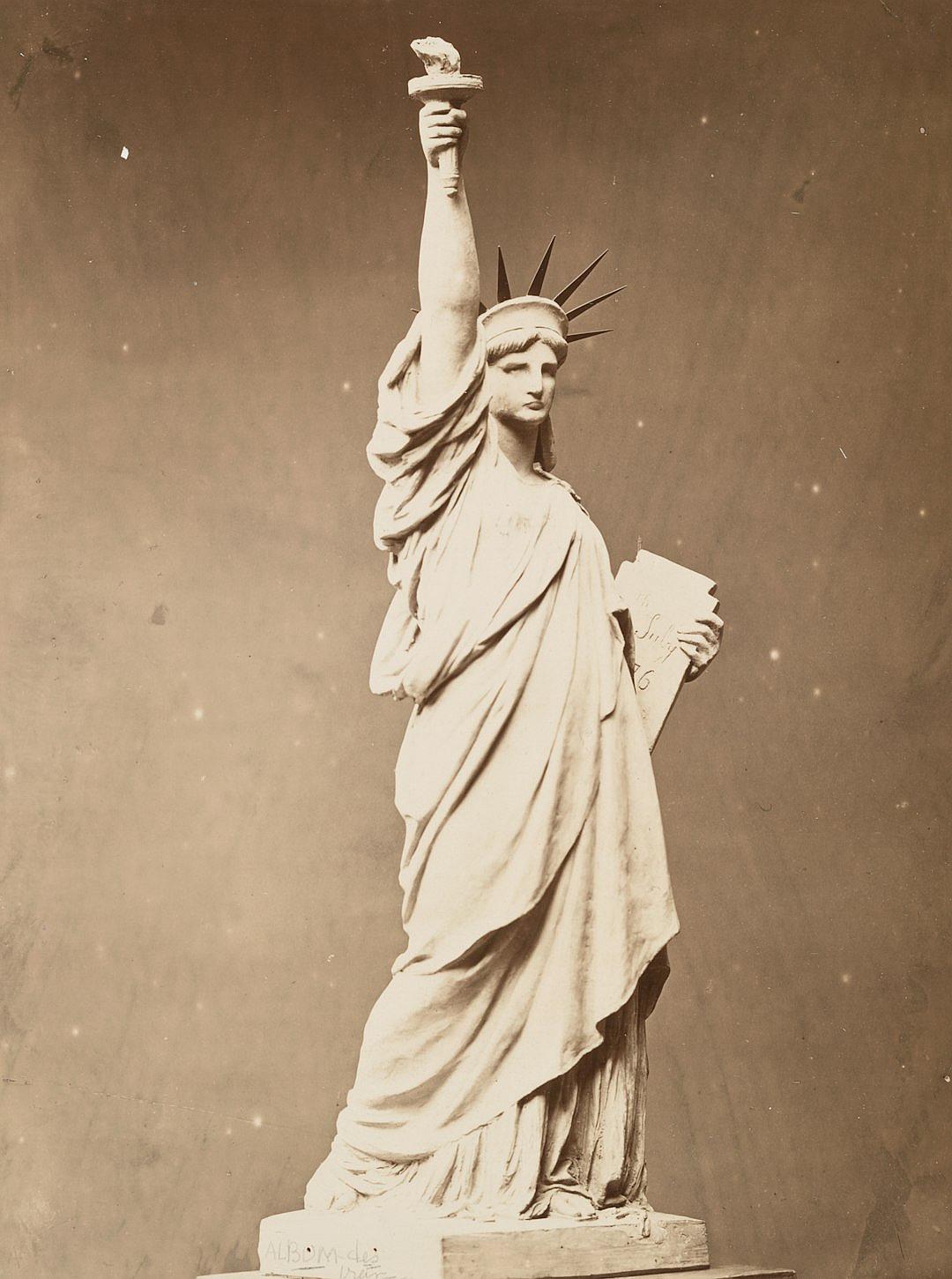 A vintage photograph of the Statue of Liberty as an old woman, holding up her torch and pointing at the viewer with one hand while showing off its intricate details in a sepiatoned setting. The statue is made from white marble, standing on top of a pedestal. The photograph is in the style of — s250