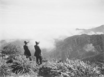 In the early morning, two men dressed in black and white suits stand on top of Mount Lushan's grassy slopes overlooking vast mountains shrouded in clouds, their figures appearing small against nature's grandeur. The scene is captured with an old-style film camera using monochrome photography, in the style of classic Hong Kong cinema aesthetics. This black-and-white photograph captures not only the scenery but also conveys historical atmosphere, evoking nostalgic memories while focusing on the men's faces.