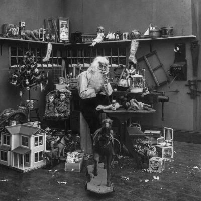 A black and white photograph of Santa Claus in his workshop, surrounded by toys like dolls, action figures, doll houses, all set against the backdrop of a simple room with wooden shelves filled with various items. The atmosphere is warm yet mysterious as he sits at an old desk, blowing into small toy horns that sit on top of each shelf. A broken rocking horse lies scattered around him, adding to the whimsical ambiance of Christmas magic., focus on face