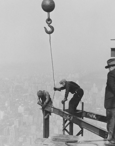 A black and white photo of construction workers on the Empire State Building, with one worker hanging from an iron hook while another watches him. The city skyline is visible in the background. The photo appears to be in the style of [Lewis Hine](https://goo.gl/search?artist%20Lewis%20Hine), a photographer known for documenting workers and social issues in the early 20th century.