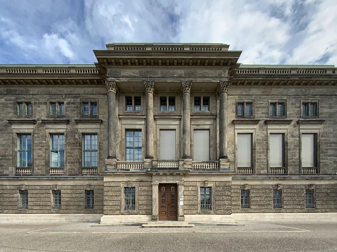 A wide shot of the front facade of an old palace in Berlin, with columns and historical details, stone walls, windows with white shades, concrete floor, front door, exterior photography in daylight under a cloudy sky with blue tones, symmetrical composition in the architectural style with detailed architectural rendering.