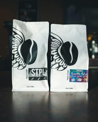 two white coffee bags with black lettering that say "STrock" and have the St_gPalmer logo on them, one bag is filled with two cargo of raw dark espresso beans from guMetroil divine t raining rain for demons in." The other has an organic label and says 'Amanana AmFeaturestoneo Malatap purely coffee beans from metallic rainbow wings. On top there's some text reading, add some watercolor, and add brand logos to each package. In the background is a bar table with moody lighting.