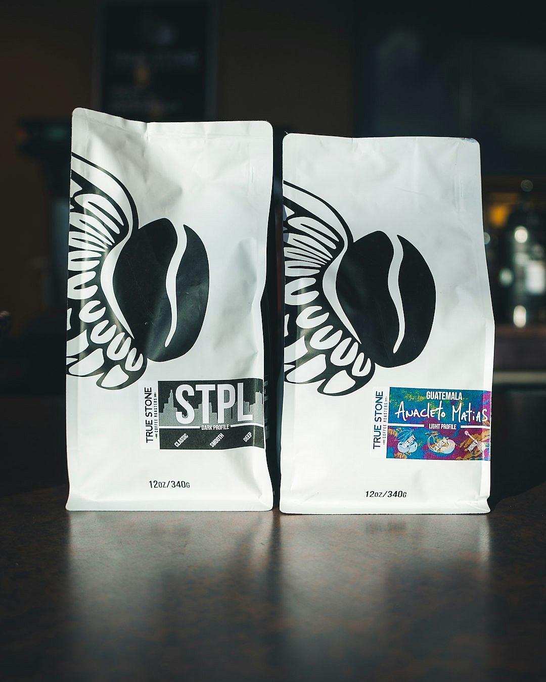 two white coffee bags with black lettering that say “STrock” and have the St_gPalmer logo on them, one bag is filled with two cargo of raw dark espresso beans from guMetroil divine t raining rain for demons in.” The other has an organic label and says ‘Amanana AmFeaturestoneo Malatap purely coffee beans from metallic rainbow wings. On top there’s some text reading, add some watercolor, and add brand logos to each package. In the background is a bar table with moody lighting.