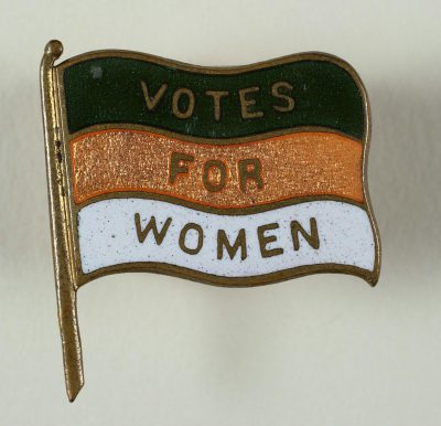 A pin with the text "VOTE FOR WOMEN" on it, in green, white and orange colors, set against an Irish flag background. It is made of enamel and gold metal. The style is vintage, reminiscent of mid 20th century writing and design, in the style of that era.