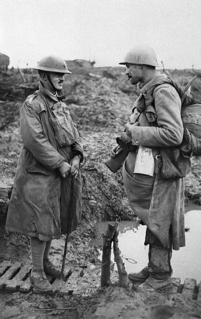 Two British soldiers in World War I, one with an overcoat and the other without, stand on muddy ground near craters of shellshuck hairs as they chat about their day's work. The soldier to his right has not taken off its helmet but is wearing it under its coat and holding two sticks that could be used for carrying bodybags or water jug by slings being worn across chest. A piece of paper hangs from around neck onto left side of jacket. black & white photo