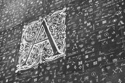 A black and white chalk drawing of a beautiful calligraphy style logo "A" on top, with lots of tiny text written all over it, on an old wall. The drawing was done in the style of calligraphy.