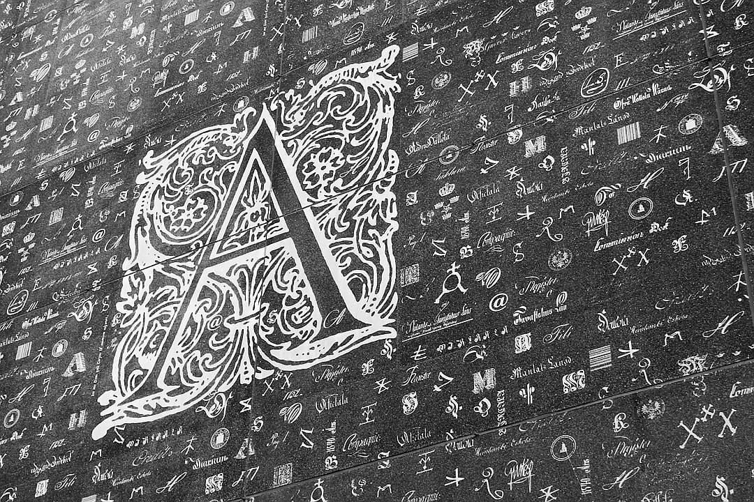 A black and white chalk drawing of a beautiful calligraphy style logo “A” on top, with lots of tiny text written all over it, on an old wall. The drawing was done in the style of calligraphy.