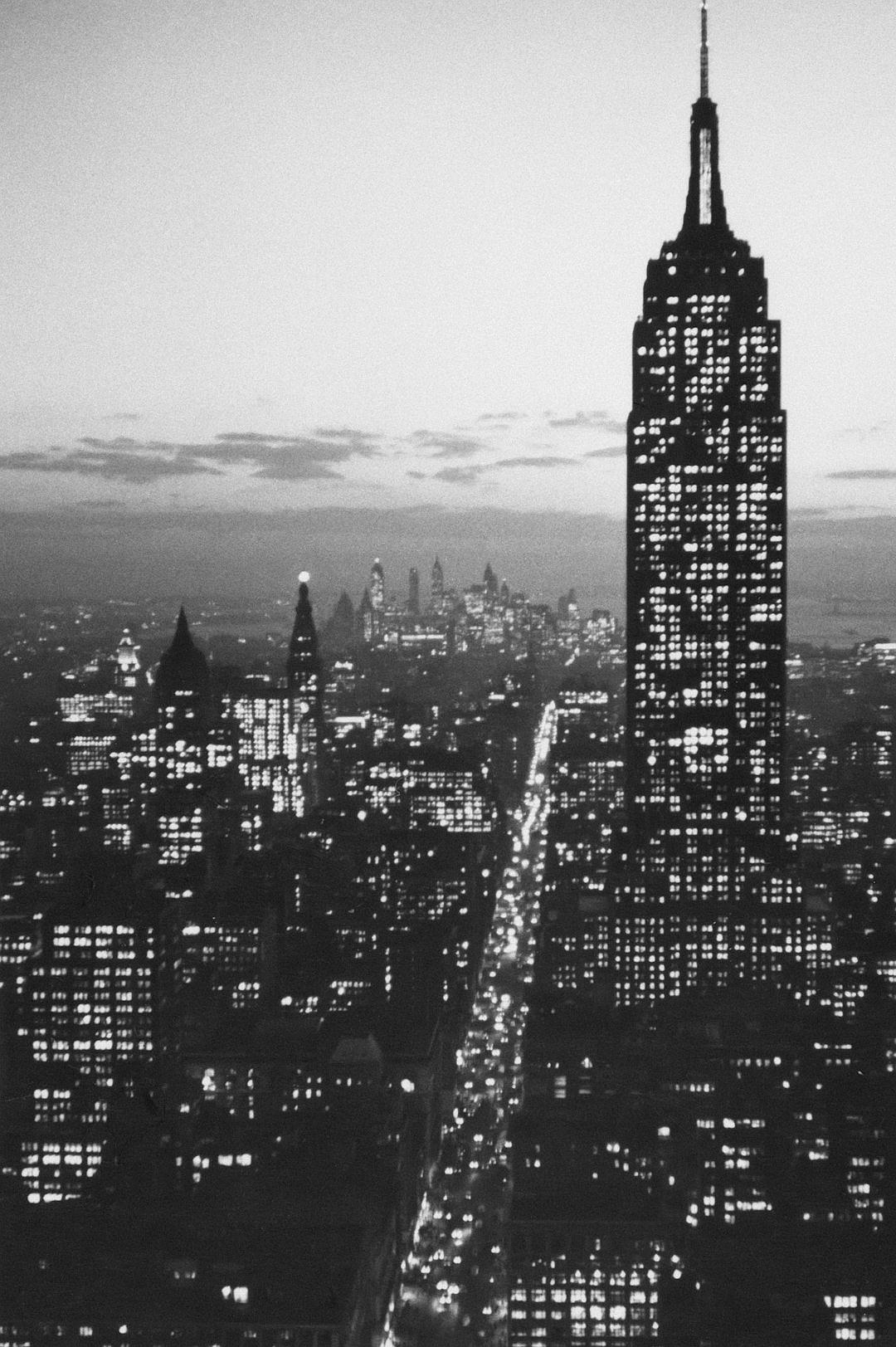 A black and white photograph of the Empire State Building at night, seen from above with buildings around it lit up in various shades of gray, capturing its iconic silhouette against an urban skyline. The city lights below create long shadows that add depth to the scene. Taken with a Black & White Film camera and wideangle lens, the 35mm film grain adds texture. High contrast lighting adds texture to details like skyscrapers and streetlights, creating a classic New York evening ambiance. The photograph is in the style of a photograph taken on 35mm film with a wideangle lens.