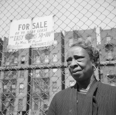 Black and white photo of a portrait of an African American woman in her mid-30s standing next to a chain link fence with a sign on it that says "Lewis & Wryther" for sale. A Third Year scourge is visible in the background. The photo was taken with a Leica M2 camera and a SummiluxMil lens in the style of the artist.