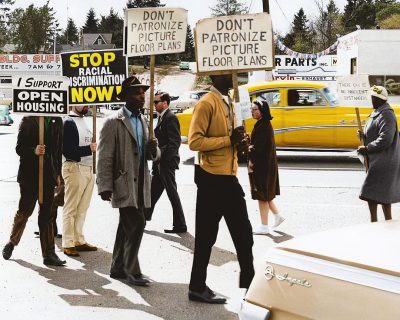 A group of African American civil rights protesters holding signs that say "DON'T ORGANIZE" and "OPEN NOW", walking in the street holding picket signs on each side of their simple yellow vintage car. They all wear hats and suit jackets. The background is an old American town, with people passing by in cars or as pedestrians carrying coffee to go. One sign reads 'ICA CENTER' , and another says 'EXIT'. The photo was taken with natural light and high resolution detail from a Sony Alpha a7 III camera, in the style of a vintage photograph.