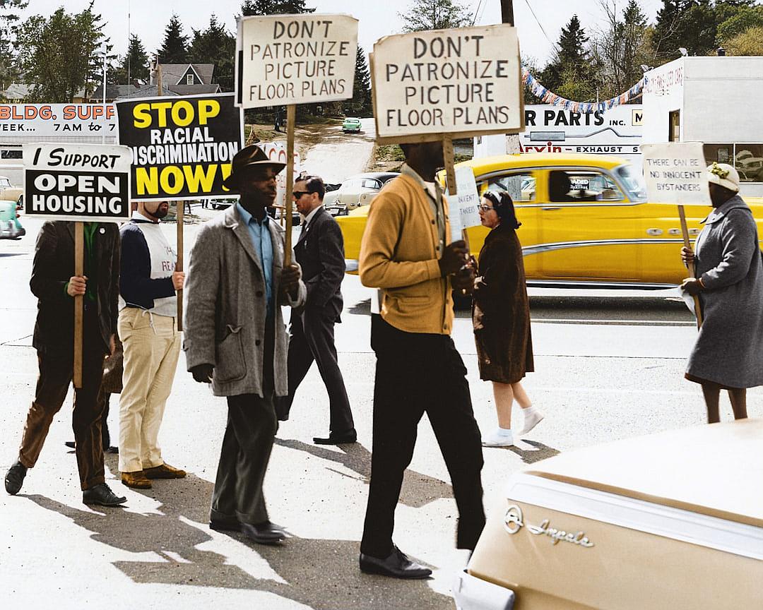 A group of African American civil rights protesters holding signs that say “DON’T ORGANIZE” and “OPEN NOW”, walking in the street holding picket signs on each side of their simple yellow vintage car. They all wear hats and suit jackets. The background is an old American town, with people passing by in cars or as pedestrians carrying coffee to go. One sign reads ‘ICA CENTER’ , and another says ‘EXIT’. The photo was taken with natural light and high resolution detail from a Sony Alpha a7 III camera, in the style of a vintage photograph.