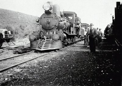 An old black and white photo of a steam engine train on railroad tracks with passengers standing next to it, in South America. The photo is in the style of an early 20th century photographer documenting travel at that time.