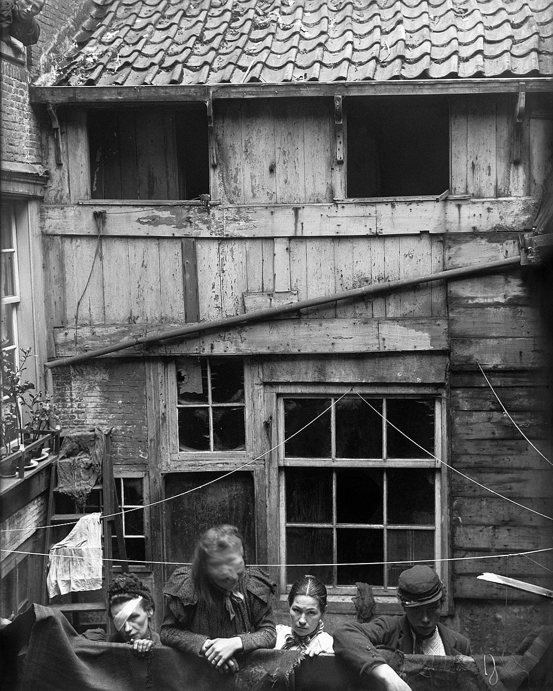 black and white photograph of poor people sitting in the backyard, looking out from behind their meat hook, wooden house with black windows, slanted roof tiles above them, in Antwerp, in the style of Swoods family style architecture, 20th century, low angle view.