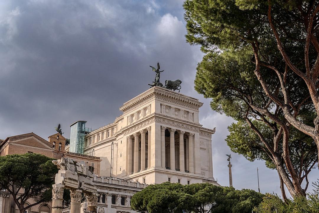 The imposing white building of the National Costa stood upright in Rome, Italy with dark clouds above it. The distant view included trees and other surrounding buildings. Shot on Canon EOS R5 camera with an aperture setting of f/8 to capture details.