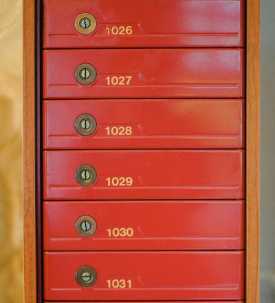 A red letterbox with six different numbers written on it, including "027" and the number of home "49". The box is located in an apartment building or community office area. There's no keyhole for each drawer. Each ripple has its own golden lock. In closeup shots, there was light coming from above, illuminating all seven drawers of the small door frame. This photo showcases the detailed view of the letters Mendes Love at sunset.