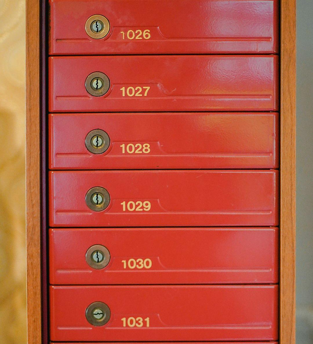 A red letterbox with six different numbers written on it, including “027” and the number of home “49”. The box is located in an apartment building or community office area. There’s no keyhole for each drawer. Each ripple has its own golden lock. In closeup shots, there was light coming from above, illuminating all seven drawers of the small door frame. This photo showcases the detailed view of the letters Mendes Love at sunset.