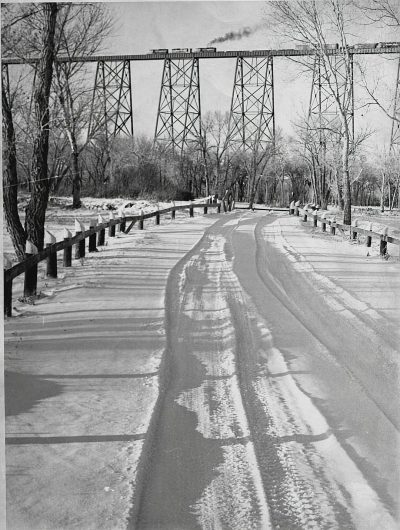 A black and white photograph of the "Big Stone Bridge" in early spring, with snow on the ground, an old train trestle over a river, trees without leaves, a road with distinct car tracks visible, snow-covered grass beside a path, distant figures walking along a street, with a vintage feel, a clear sky, early morning light casting long shadows, a nostalgic atmosphere.