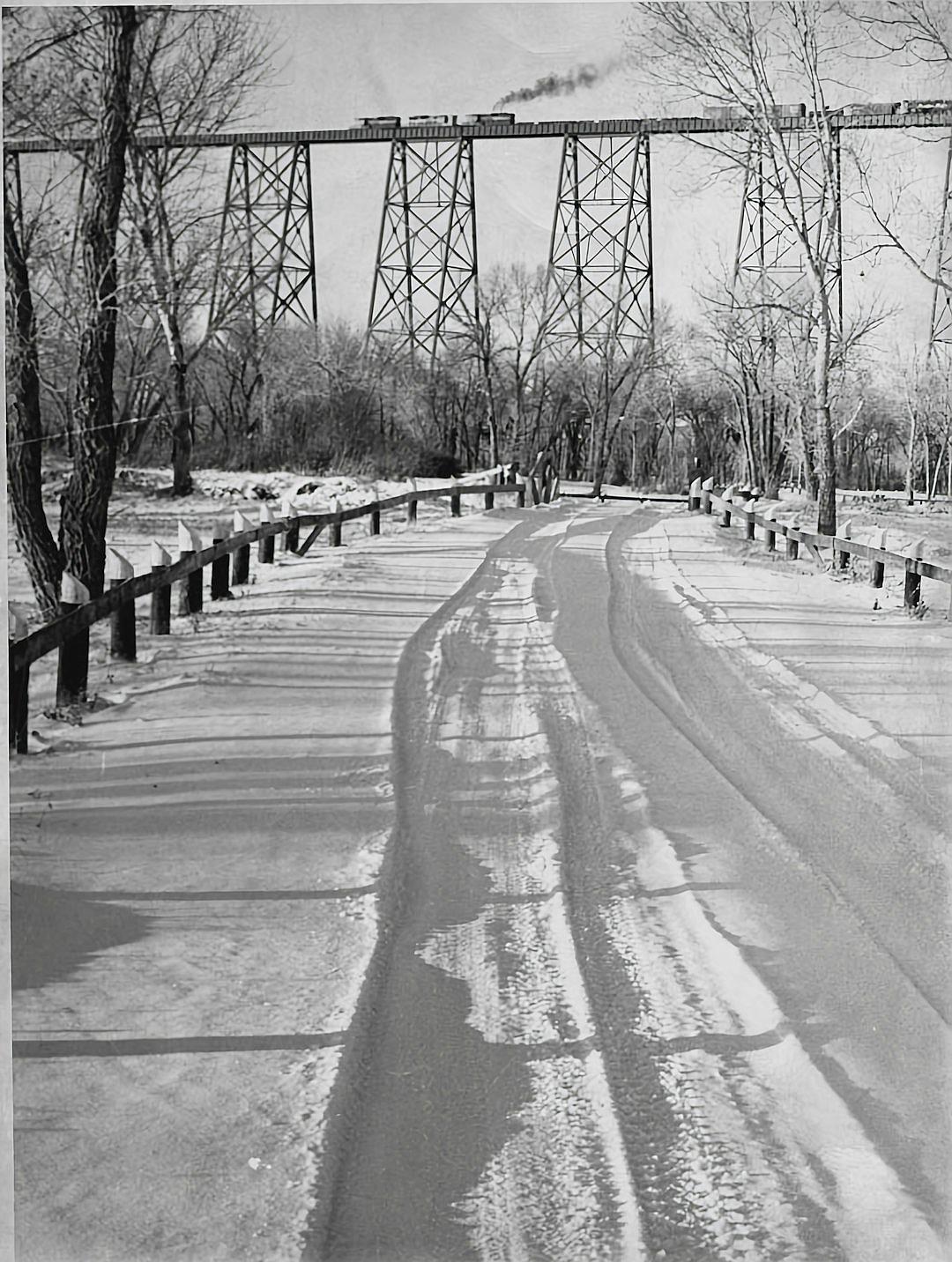 A black and white photograph of the “Big Stone Bridge” in early spring, with snow on the ground, an old train trestle over a river, trees without leaves, a road with distinct car tracks visible, snow-covered grass beside a path, distant figures walking along a street, with a vintage feel, a clear sky, early morning light casting long shadows, a nostalgic atmosphere.