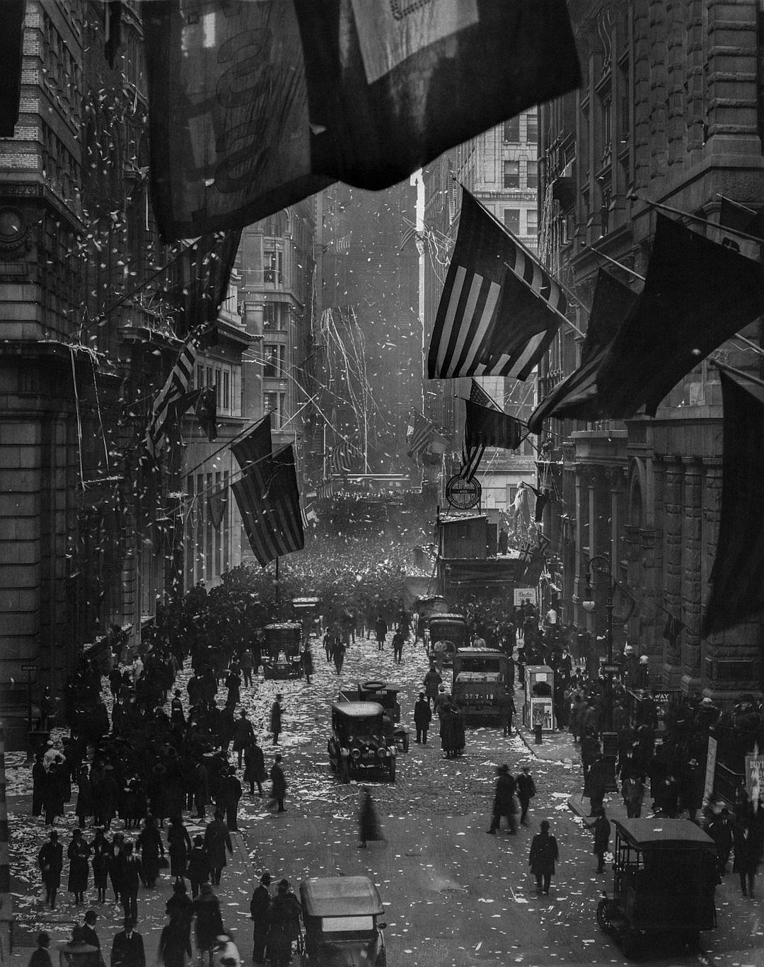 A black and white photograph of the Great Wall Street in New York City, showing people celebrating with flags, confetti, streetlights, horse-drawn carts and public transportation, as well as buildings decorated for Christmas. The photo was taken in the style of John Ro Martin using an early 20th century camera, capturing intricate details of urban life during that era.