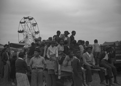 A black and white photograph of young African American men, women and children sitting on the back of a flatbed truck at an old carnival in rural southwest Louisiana with a ferris wheel in the background. They all wear simple  and some have hats or bow ties or plaid shirts. The photo was shot from behind them. The sky is overcast. This photo was taken in the style of [Dorothea Lange](https://goo.gl/search?artist%20Dorothea%20Lange).