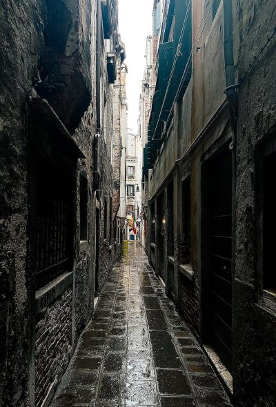 A photo of an alleyway in Venice, Italy on a rainy day. The narrow street is lined with ancient buildings and the ground reflects wet black stone tiles. A person can be seen walking down one side of it under cover from the rain in the style of nearby building eaves. It's a moody scene with grey clouds overhead and a sense of solitude in its quietness.