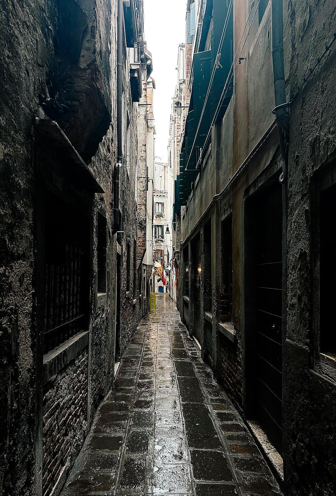 A photo of an alleyway in Venice, Italy on a rainy day. The narrow street is lined with ancient buildings and the ground reflects wet black stone tiles. A person can be seen walking down one side of it under cover from the rain in the style of nearby building eaves. It’s a moody scene with grey clouds overhead and a sense of solitude in its quietness.