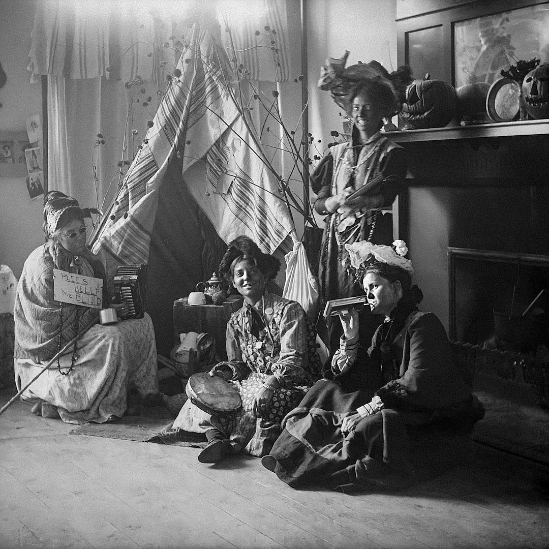 A black and white photo depicts women in fancy dress sitting on the floor around an Indian basket containing wooden sticks. They have listening devices and headphones next to them. They all look sad as if attending some kind of party where no one seems happy. The room has curtains, wallpaper, and furniture from early 20th century New York City decor including small sofas and coffee tables. There is also a wall full of framed pictures. It feels like a scene from early cinema in the style of [Edward Hopper](https://goo.gl/search?artist%20Edward%20Hopper).